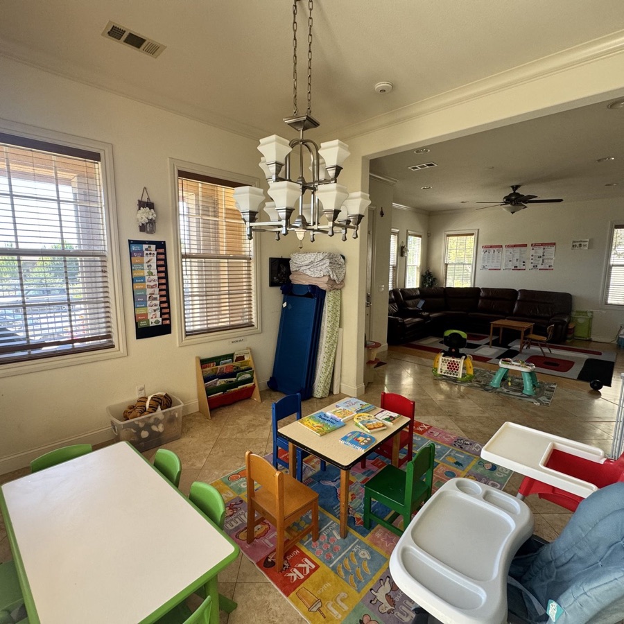 Bright warm play area at Madhu's Home Daycare — chandelier, kid-sized tables and chairs, colorful rugs, natural light through big windows