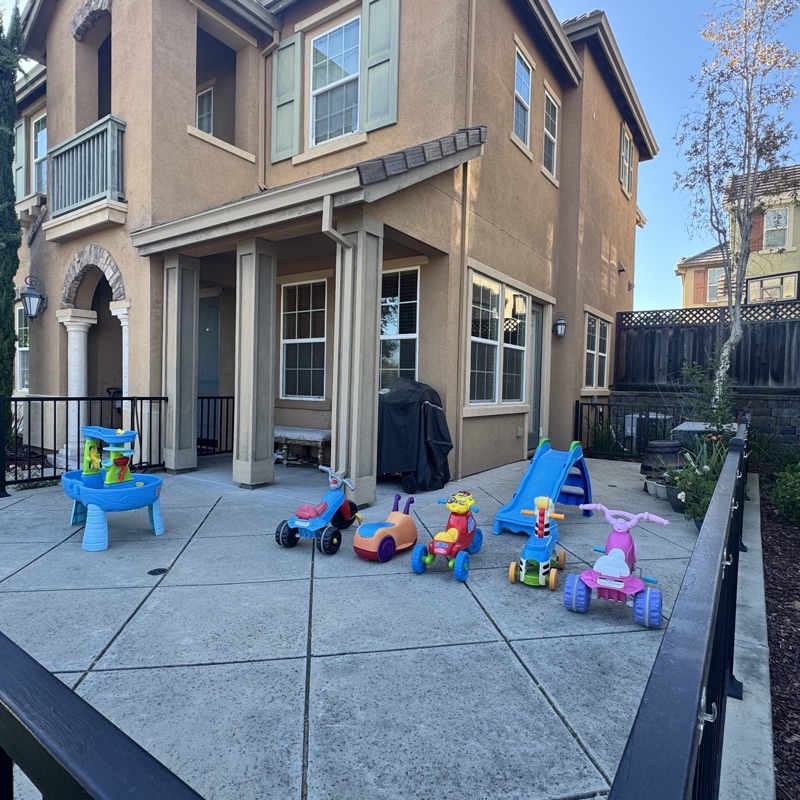 Fenced front patio with a blue slide, a water table, and colorful ride-on toys ready for outdoor play