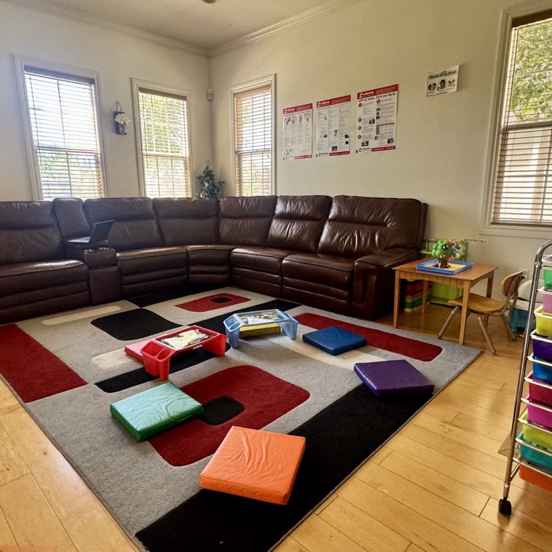Big brown leather sectional sofa with colorful floor cushions on a patterned rug — bright natural light through the windows